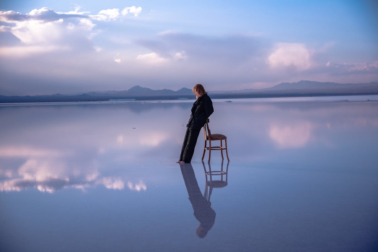 A woman stands by a chair on a serene salt flat, reflecting beautiful clouds and mountains.